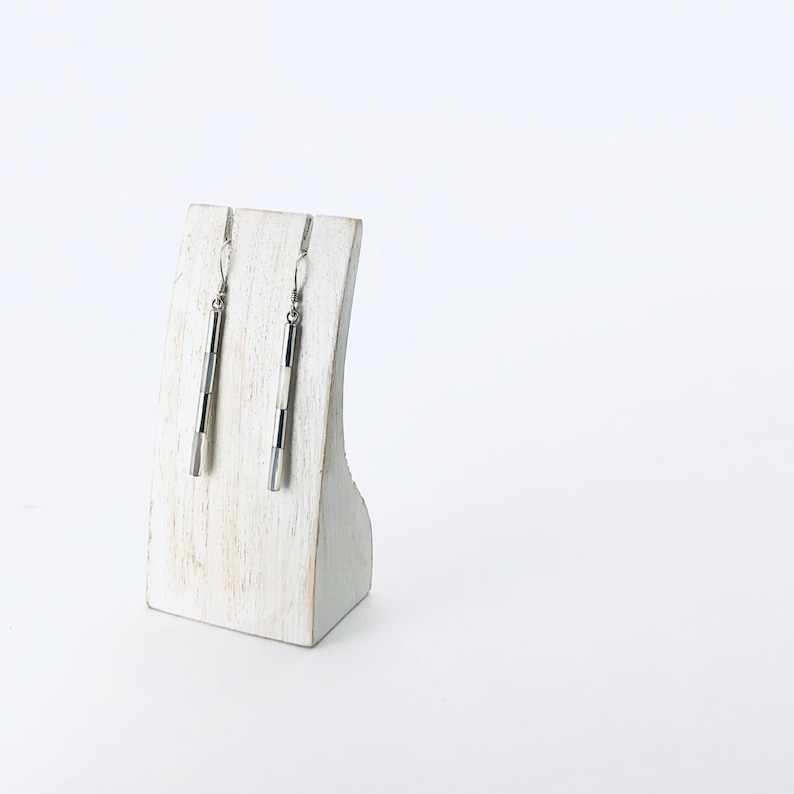 May include: A pair of silver dangle earrings with black and white geometric designs. The earrings are displayed on a white wooden stand.