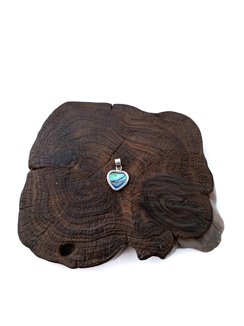 May include: A silver heart-shaped pendant with a blue and green abalone shell inlay. The pendant is displayed on a dark brown wooden surface.