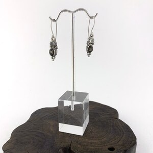 May include: Silver dangle earrings with a black and white stone design. The earrings are displayed on a wooden stand.