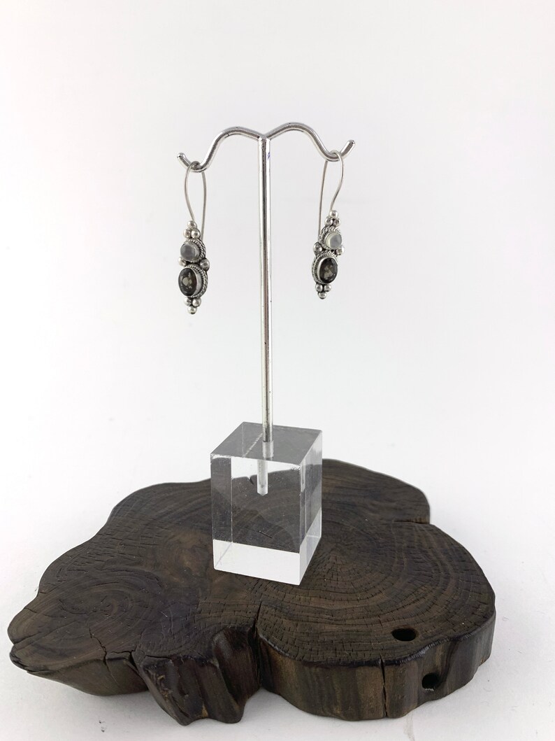 May include: Silver dangle earrings with a black and white stone design. The earrings are displayed on a wooden stand.