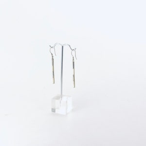 May include: A pair of silver dangle earrings with white rectangular beads. The earrings are hanging on a silver stand.