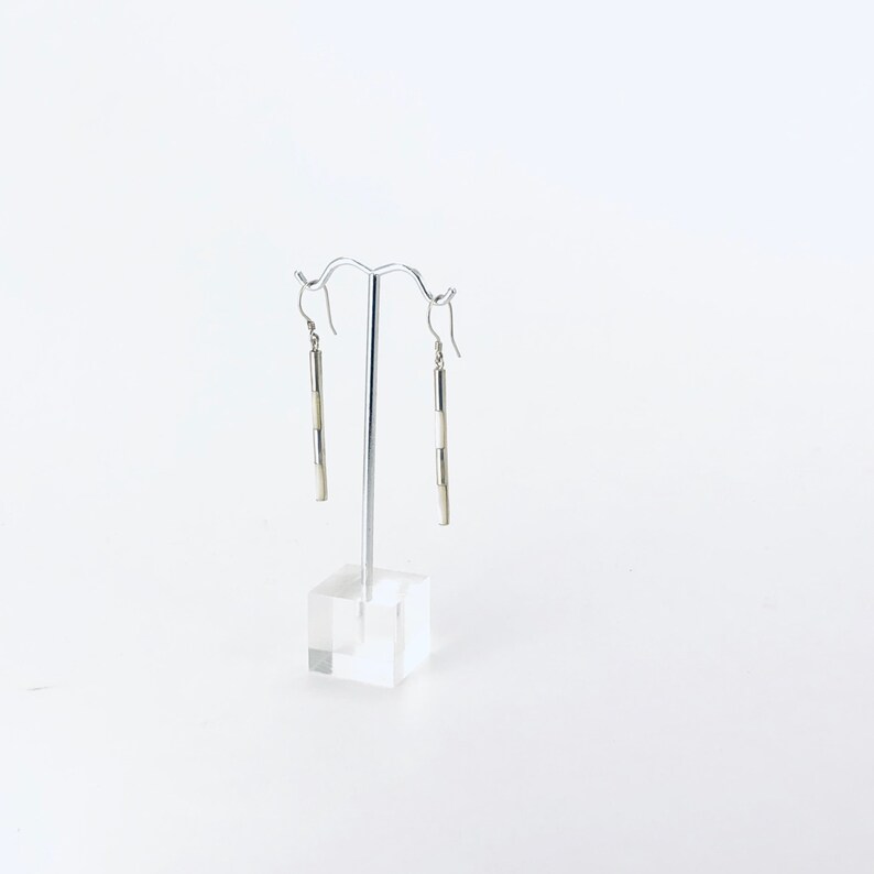 May include: A pair of silver dangle earrings with white rectangular beads. The earrings are hanging on a silver stand.
