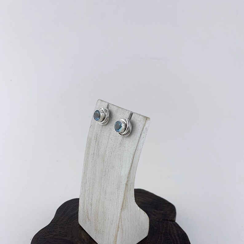 May include: A pair of silver stud earrings with blue topaz gemstones. The earrings are displayed on a white wooden stand.