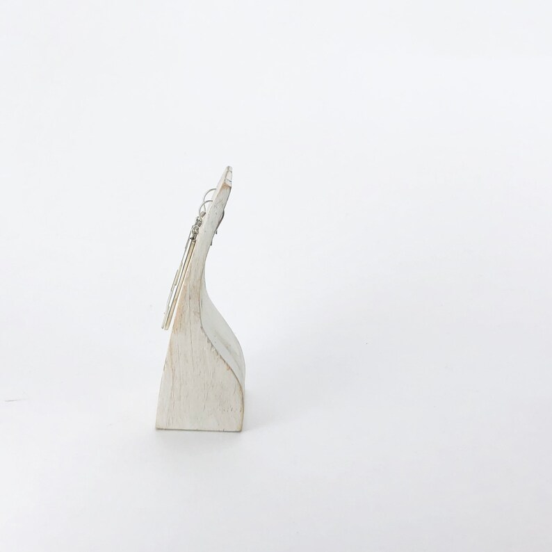 May include: Silver dangle earrings with a simple design, displayed on a white wooden stand.
