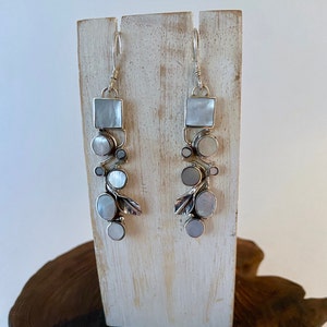 May include: A pair of silver earrings featuring white mother-of-pearl stones in a variety of shapes and sizes. The earrings are displayed on a wooden stand.