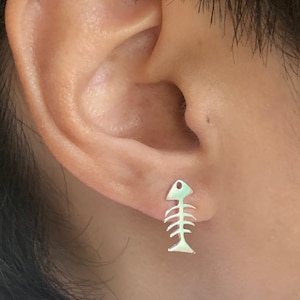 May include: Silver fishbone earrings. The earrings are in the shape of a fish skeleton. The earrings are hanging from the earlobe.