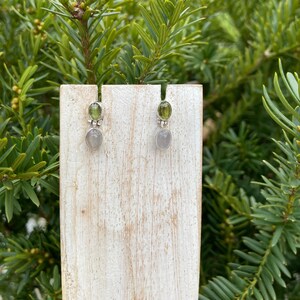 May include: A pair of silver earrings with green and grey stones. The earrings are displayed on a wooden stand.
