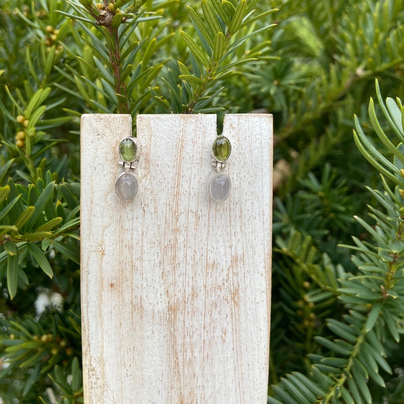 May include: A pair of silver earrings with green and grey stones. The earrings are displayed on a wooden stand.