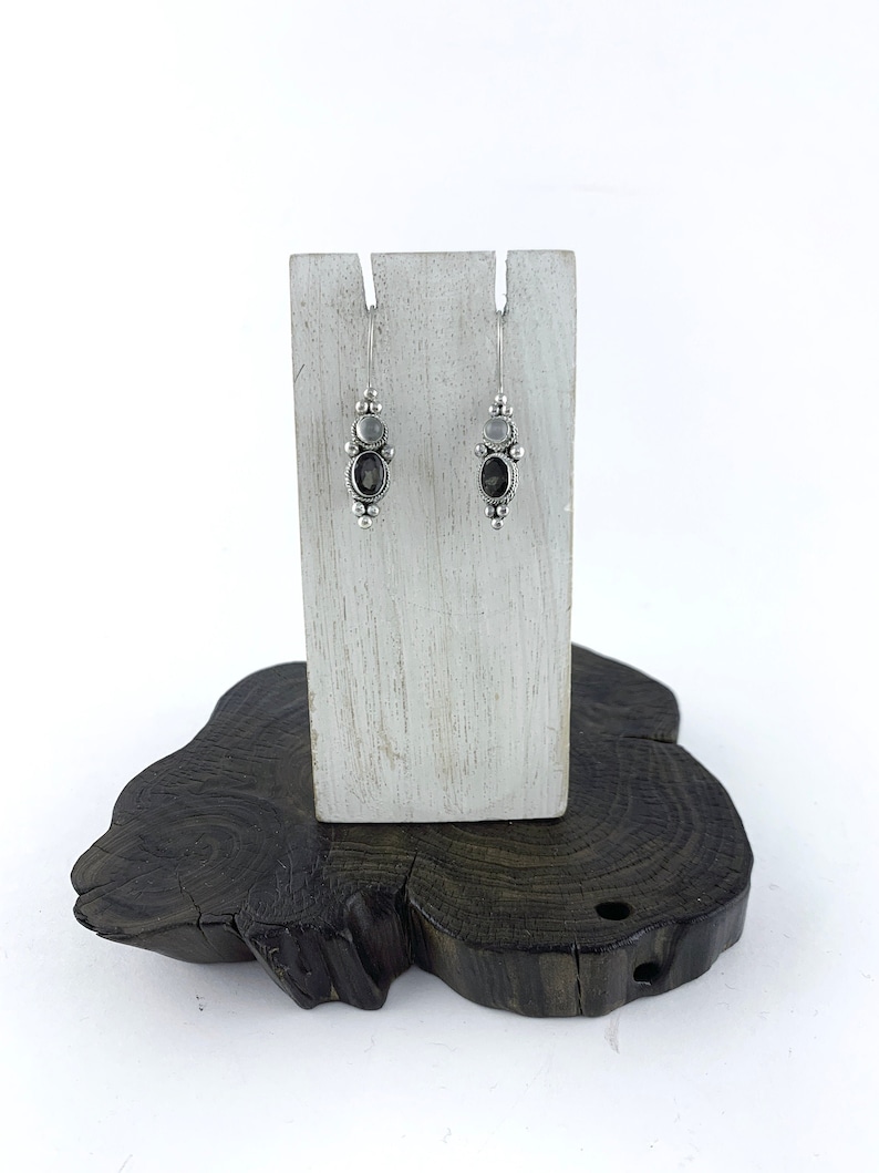 May include: A pair of silver dangle earrings with a dark gray oval gemstone and smaller round clear gemstones. The earrings are displayed on a white wooden stand.