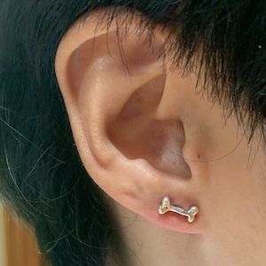 May include: A close-up of a person's ear with a small silver bone-shaped earring.