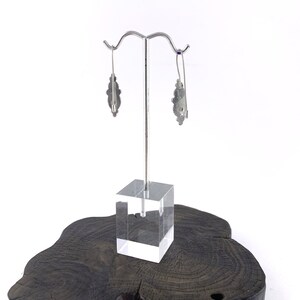 May include: A pair of silver earrings with a floral design. The earrings are hanging from a silver stand on a wooden base.