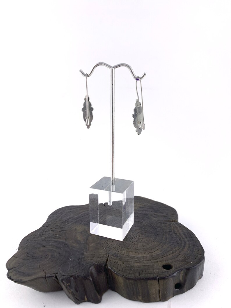 May include: A pair of silver earrings with a floral design. The earrings are hanging from a silver stand on a wooden base.