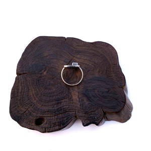 May include: A silver ring with a square-shaped stone set in the center, displayed on a dark brown wooden surface.