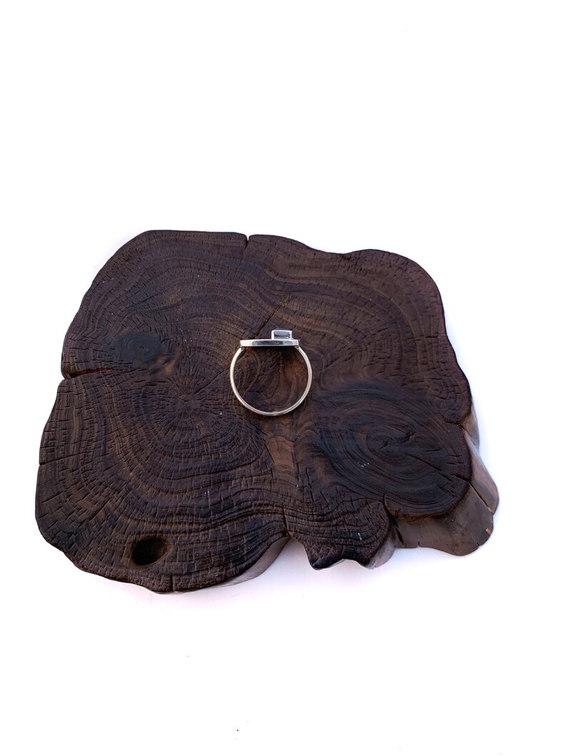 May include: A silver ring with a square-shaped stone set in the center, displayed on a dark brown wooden surface.
