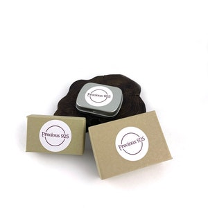 May include: Three small boxes with the text "Precious 925" on them. The boxes are brown and white and are sitting on a wooden surface.
