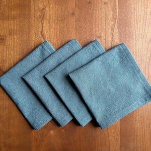 May include: Four square, denim-colored cloth napkins are arranged diagonally on a wooden surface. The napkins are a muted blue-gray color and appear to be made of a textured fabric. The wooden surface has a warm, reddish-brown tone.