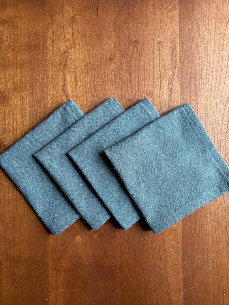 May include: Four square, denim-colored cloth napkins are arranged diagonally on a wooden surface. The napkins are a muted blue-gray color and appear to be made of a textured fabric. The wooden surface has a warm, reddish-brown tone.