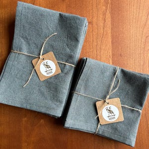 May include: Two stacks of folded, gray linen napkins tied with twine. Each stack has a small, square tag with a heron illustration and the word "HERON". The napkins are on a wooden surface.