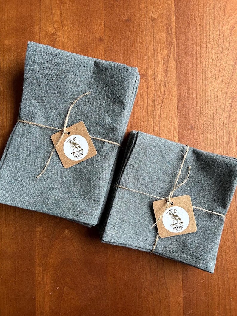 May include: Two stacks of folded, gray linen napkins tied with twine. Each stack has a small, square tag with a heron illustration and the word "HERON". The napkins are on a wooden surface.