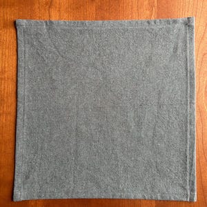 May include: A rectangular, gray linen napkin with a stitched border. The napkin is displayed on a wooden surface, showcasing its simple design and texture. The fabric appears to be a medium weight.