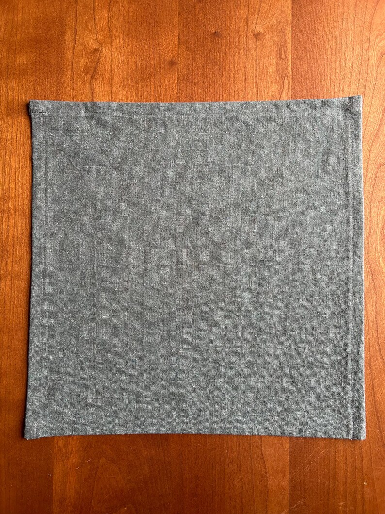 May include: A rectangular, gray linen napkin with a stitched border. The napkin is displayed on a wooden surface, showcasing its simple design and texture. The fabric appears to be a medium weight.