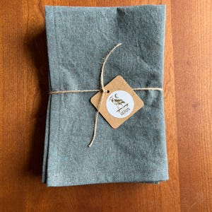 May include: A folded, teal-colored linen cloth, tied with twine and a small, brown tag. The tag features a white circle with a heron illustration and the word "HERON". The cloth is neatly stacked, suggesting a set of napkins or towels.