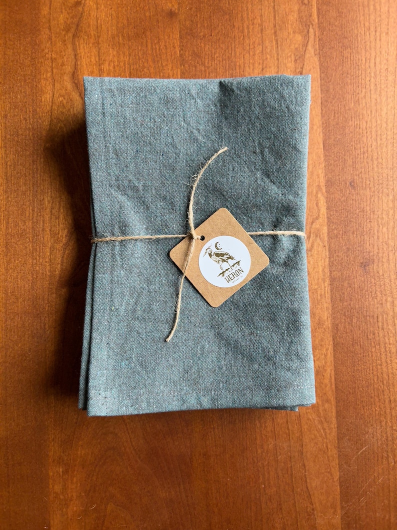 May include: A folded, teal-colored linen cloth, tied with twine and a small, brown tag. The tag features a white circle with a heron illustration and the word "HERON". The cloth is neatly stacked, suggesting a set of napkins or towels.