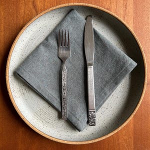 May include: A place setting featuring a gray speckled ceramic plate with a gray linen napkin. A silver fork with a decorative handle and a silver knife are placed on the napkin. The setting is on a wooden surface.