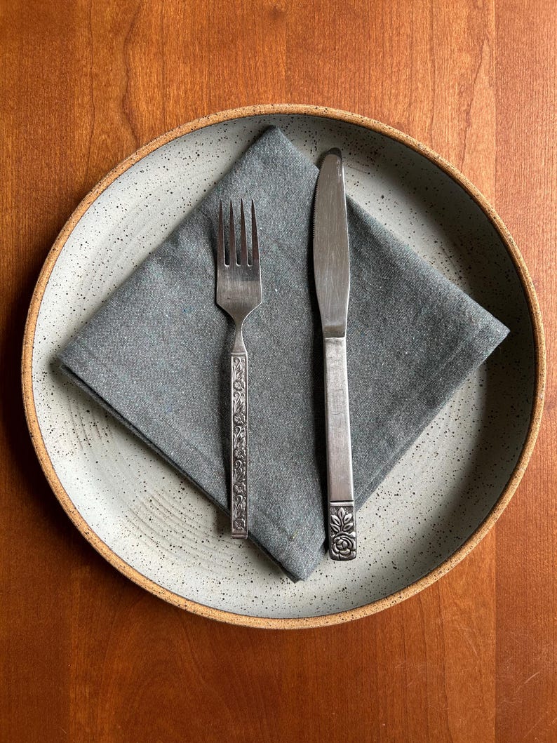 May include: A place setting featuring a gray speckled ceramic plate with a gray linen napkin. A silver fork with a decorative handle and a silver knife are placed on the napkin. The setting is on a wooden surface.