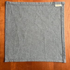 May include: A square, gray linen napkin with a stitched border. The napkin has a small rectangular tag in the upper right corner with the word "HERON" printed on it. The napkin is displayed on a wooden surface.
