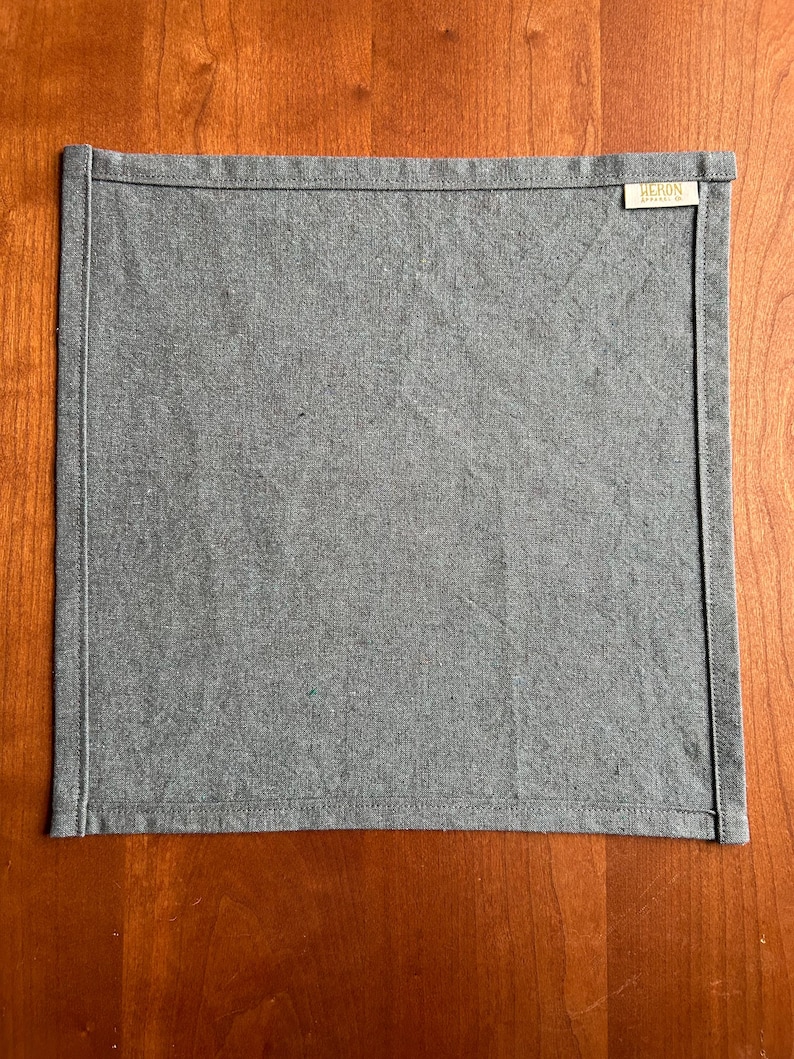May include: A square, gray linen napkin with a stitched border. The napkin has a small rectangular tag in the upper right corner with the word "HERON" printed on it. The napkin is displayed on a wooden surface.
