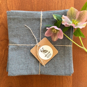 May include: A stack of folded, gray linen napkins tied with twine, featuring a small, brown tag with a heron illustration and the word "HERON". Pink and yellow flowers with green leaves are placed next to the napkins.