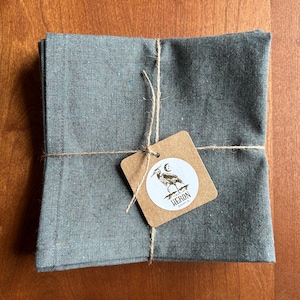 May include: A stack of folded, gray-green linen napkins tied with twine. A small, brown tag is attached, featuring a heron illustration and the word "HERON". The napkins are neatly arranged on a wooden surface.