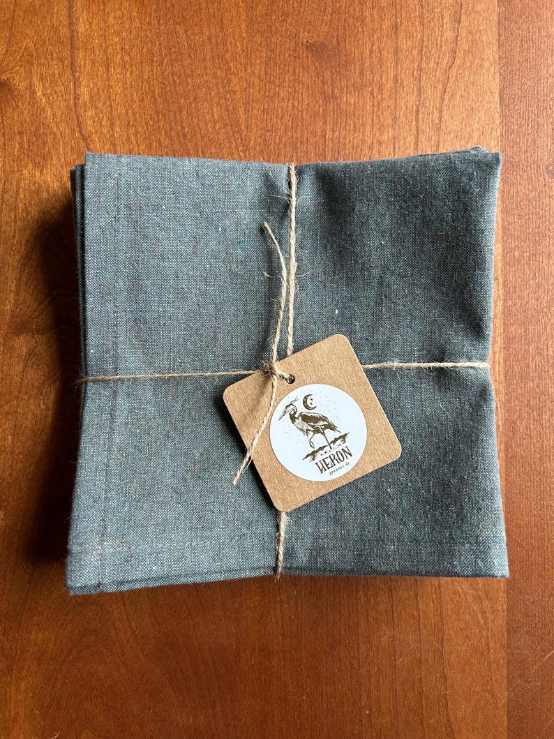 May include: A stack of folded, gray-green linen napkins tied with twine. A small, brown tag is attached, featuring a heron illustration and the word "HERON". The napkins are neatly arranged on a wooden surface.