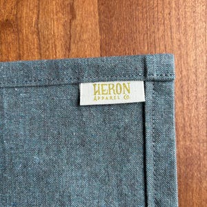 May include: Close-up of a textured, gray-blue fabric with a stitched edge. A rectangular label with the text "HERON APPAREL CO." in gold lettering is sewn onto the fabric. The background is a wooden surface.