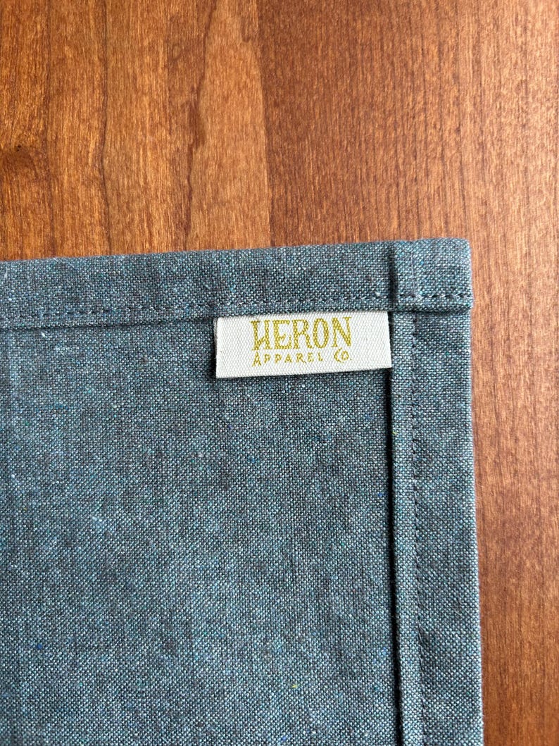 May include: Close-up of a textured, gray-blue fabric with a stitched edge. A rectangular label with the text "HERON APPAREL CO." in gold lettering is sewn onto the fabric. The background is a wooden surface.