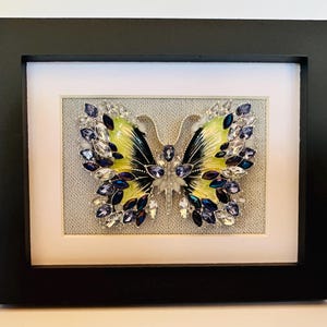 May include: Framed butterfly art piece. The butterfly has yellow and black wings, adorned with blue and clear crystals. The artwork is set against a light gray background within a white mat and a black frame.