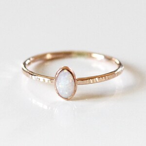 May include: A rose gold ring featuring a small, oval-shaped white opal stone set in a bezel. The thin band of the ring has a textured, hammered finish that catches the light, creating a subtle, reflective surface across the metal band.
