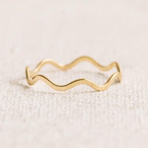 May include: A delicate gold ring with a wavy, undulating design. The ring's surface reflects light, highlighting its smooth, polished finish. The ring is a minimalist piece of jewelry.