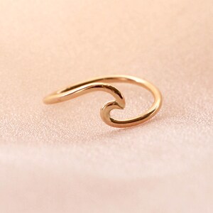 May include: A gold-coloured ring shaped like a wave. The ring has a simple, elegant design with a smooth, polished finish. The wave design is a popular symbol of the ocean and surfing.