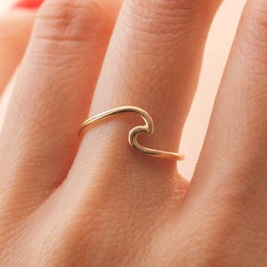 May include: A gold-coloured ring in the shape of a wave. The ring is worn on a finger, with the wave design facing upwards. The ring appears to be made of a thin metal band.