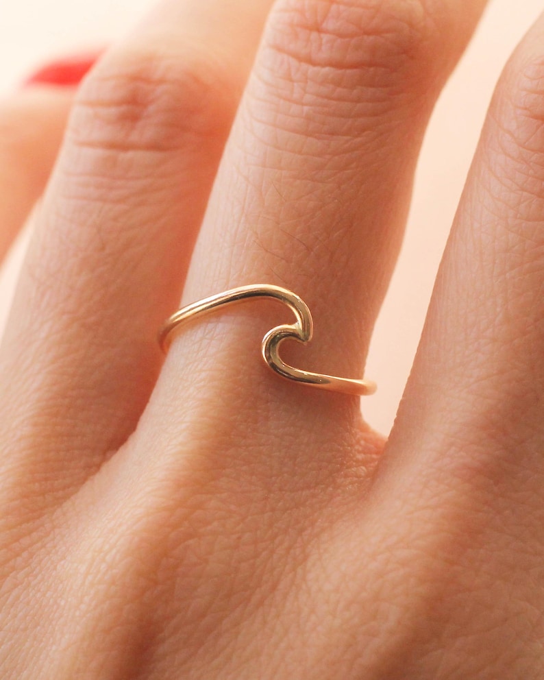 May include: A gold-coloured ring in the shape of a wave. The ring is worn on a finger, with the wave design facing upwards. The ring appears to be made of a thin metal band.