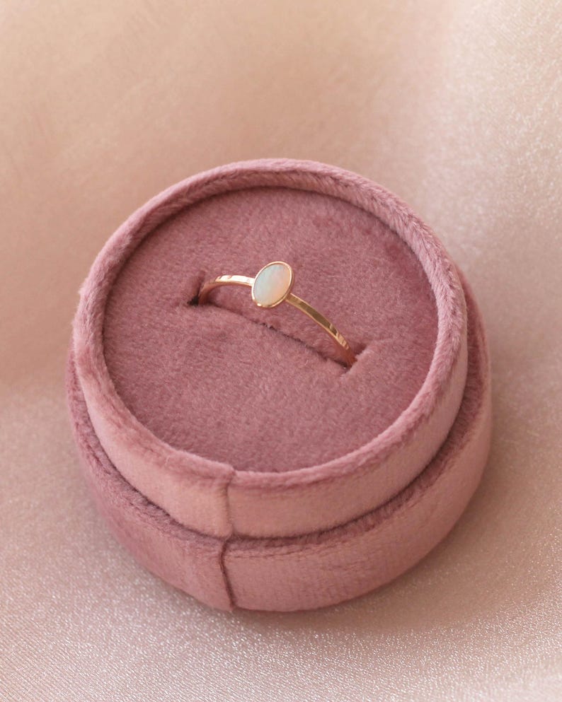 May include: A thin gold ring featuring a small, smooth, oval-shaped white stone set horizontally. The ring is nestled securely within the center slot of a round, dusty rose-colored velvet jewelry box with a soft, plush texture.