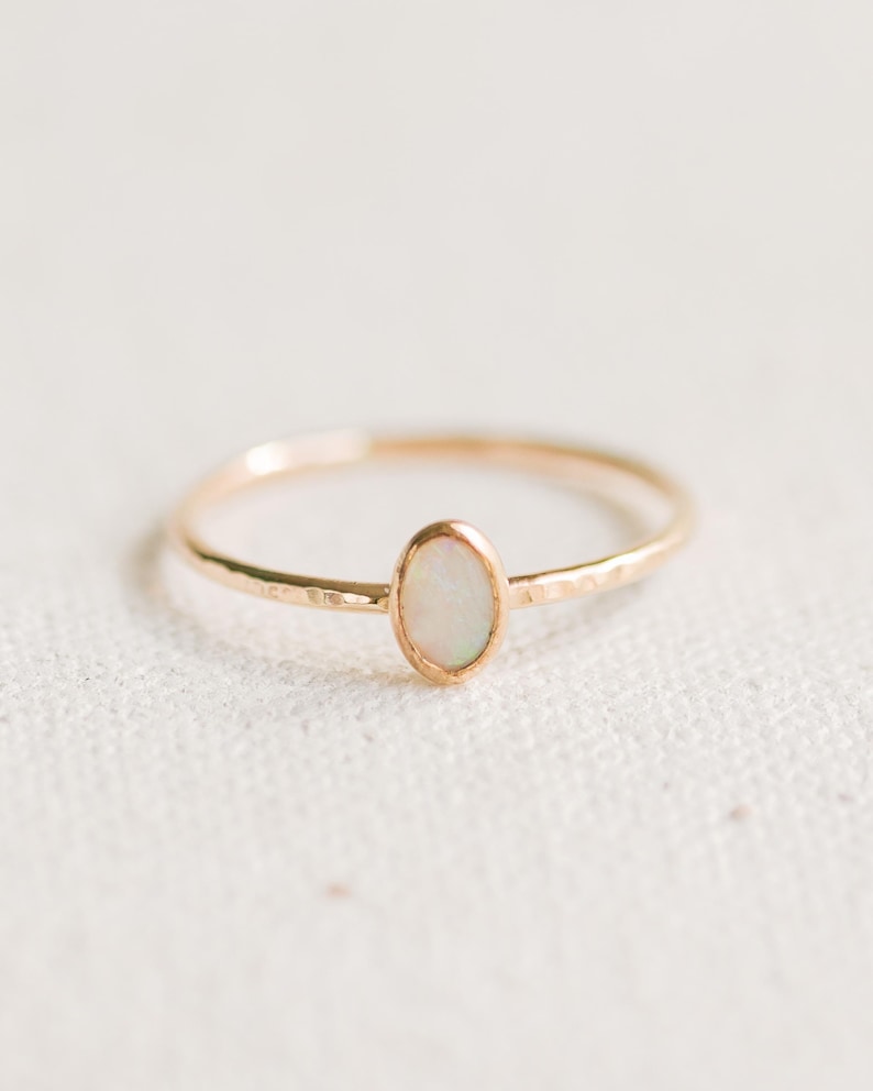 May include: A thin gold ring featuring a small, vertically oriented oval opal gemstone set in a bezel. The band of the ring has a subtle hammered texture, creating a reflective surface that catches the light along the thin, circular metal band.