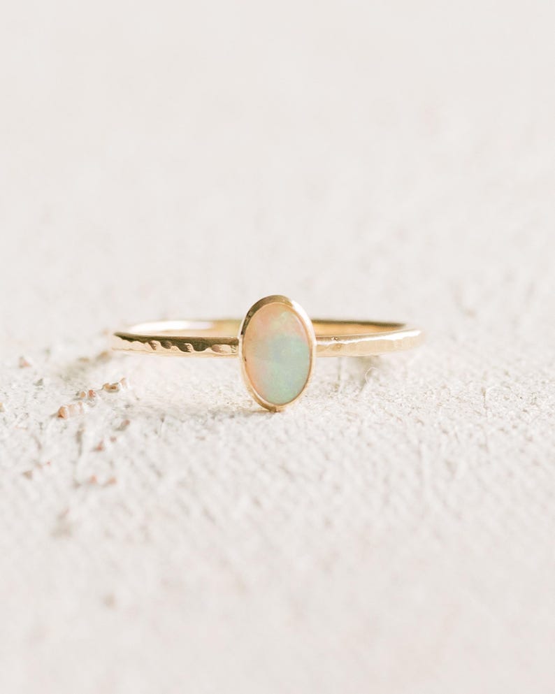 May include: A gold ring featuring a thin, hammered band and a central oval-shaped opal gemstone. The opal displays iridescent flashes of color, and the metal band has a textured, uneven surface finish throughout its circular structure.