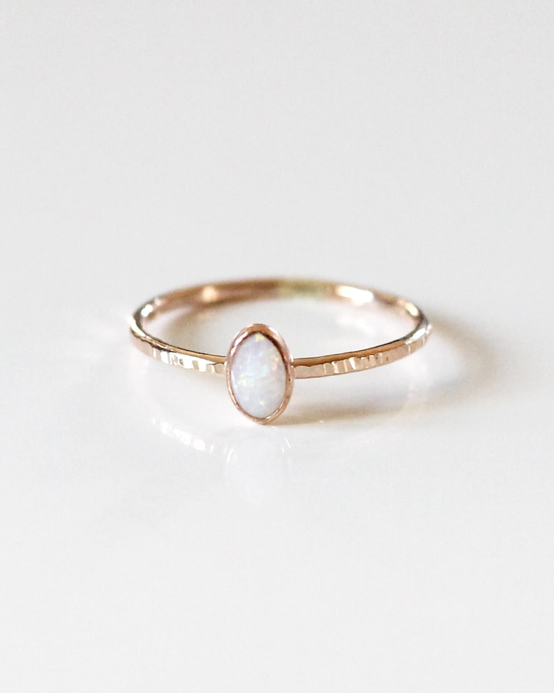 May include: A rose gold ring featuring a small, oval-shaped white opal stone set in a bezel. The thin band of the ring has a textured, hammered finish that catches the light, creating a subtle, reflective surface across the metal band.