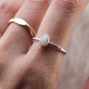 May include: A silver ring featuring a small, oval-shaped white opal gemstone set in a bezel. The band of the ring has a textured, hammered finish. A second, plain gold-colored band is visible on an adjacent finger, completing the jewelry set.