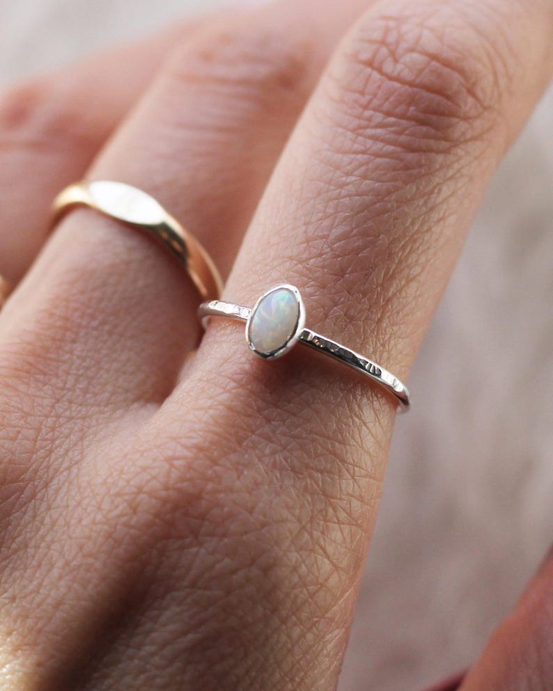 May include: A silver ring featuring a small, oval-shaped white opal gemstone set in a bezel. The band of the ring has a textured, hammered finish. A second, plain gold-colored band is visible on an adjacent finger, completing the jewelry set.
