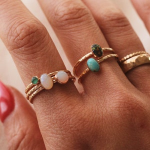 May include: A collection of gold-toned rings featuring hammered bands and various oval-shaped gemstones. The rings include small green, white, and turquoise stones, along with a dark speckled stone and a wider band with a textured, dimpled surface.