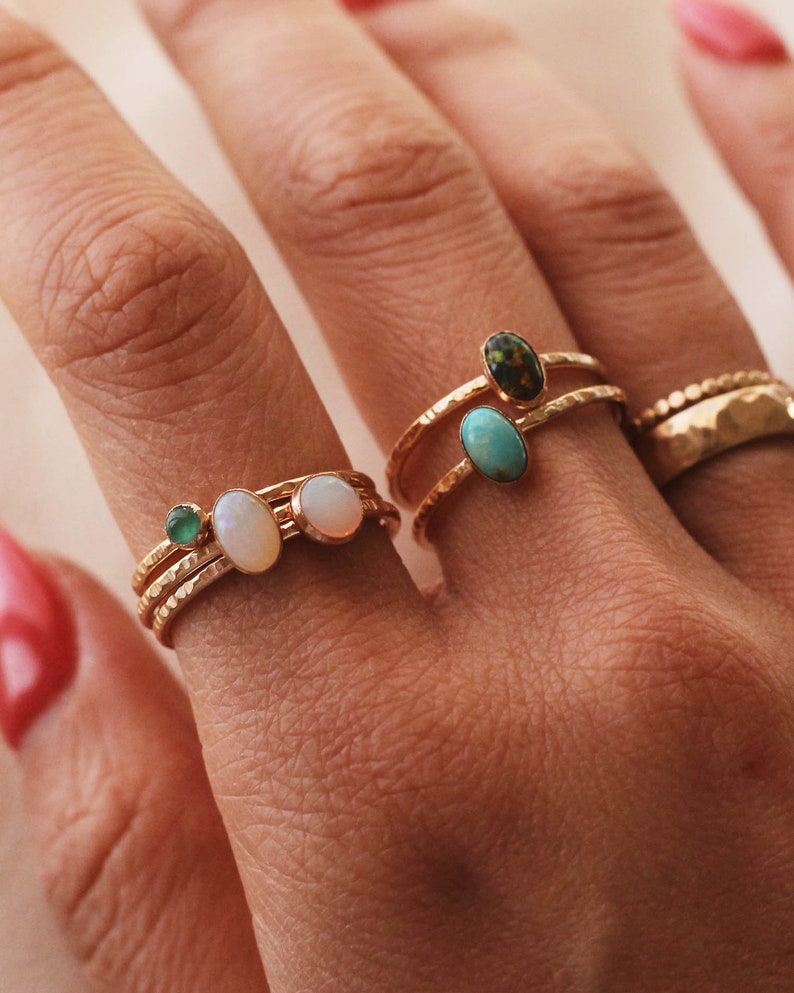 May include: A collection of gold-toned rings featuring hammered bands and various oval-shaped gemstones. The rings include small green, white, and turquoise stones, along with a dark speckled stone and a wider band with a textured, dimpled surface.